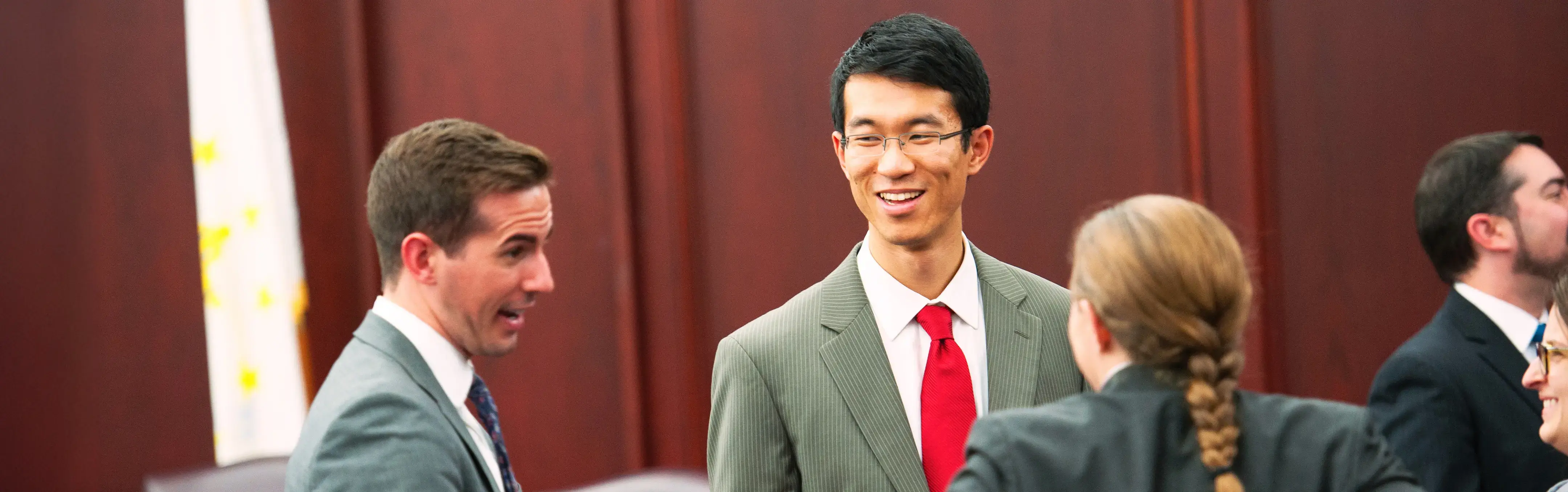 Group of professionals in suits having an animated conversation in a courtroom with wood paneling and a state flag visible. 