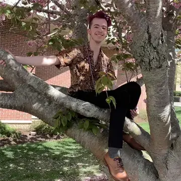 Student in a patterned shirt sits smiling on a tree branch on campus.