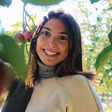 Student in a turtleneck sweater smiles outdoors among apple trees.