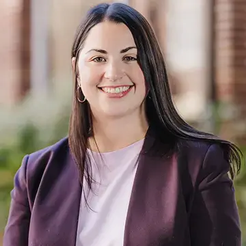 Alumna in a purple blazer and light top smiles in front of a brick building.