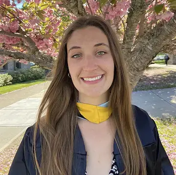 Roger Williams University graduate in cap and gown smiles outdoors beneath blooming cherry blossoms.