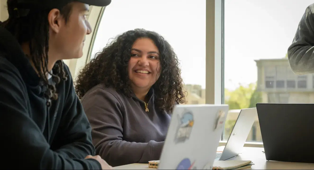 Two students smiling at each other seated in front of laptop computers